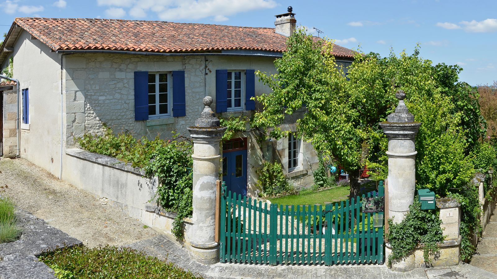 Maison en pierre aux volets bleus derrière un portail vert et de la vigne vierge