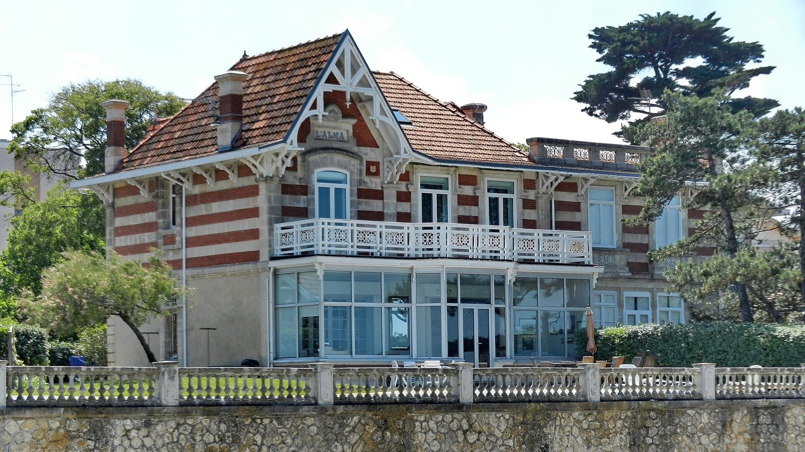Villa balnéaire en briques et pierre, avec balcon blanc, face à la mer derrière un muret